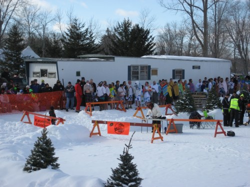 A crowd of people in winter clothing gathers near a trailer, surrounded by snow and orange safety barriers.