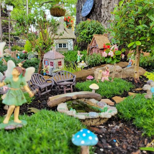 A whimsical fairy garden with miniature houses, figurines, benches, and lush greenery.
