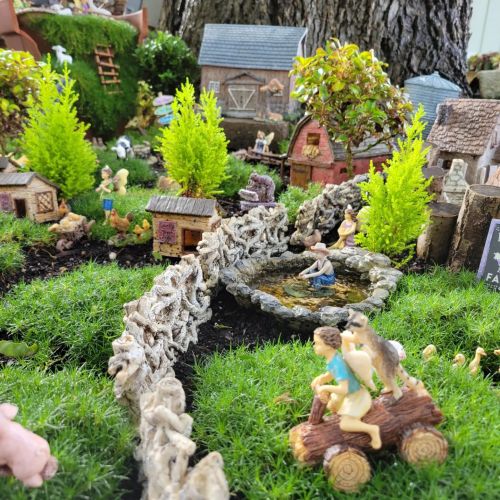 A whimsical miniature village with tiny houses, figures, and a pond, surrounded by greenery and decorative elements.