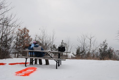 Two people sit on a picnic table in the snow, surrounded by trees, with sleds nearby and a cloudy sky above.
