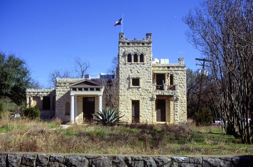 Historic stone building with a tower, Texas flag, and overgrown landscaping against a clear blue sky.