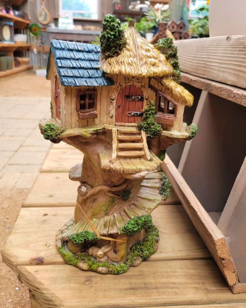 A whimsical miniature treehouse with a thatched roof, nestled on a tree stump, surrounded by greenery.