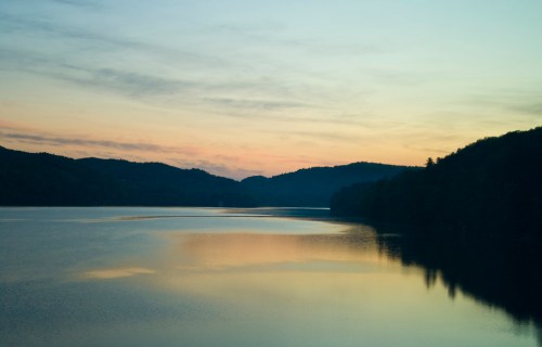 Serene lake at sunset, reflecting soft colors with mountains in the background and a calm water surface.