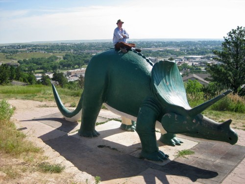 A person sits on a large green triceratops statue overlooking a scenic landscape.