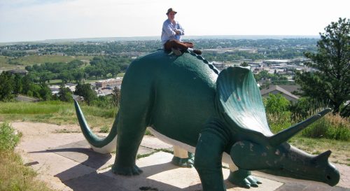 A person sits atop a large green triceratops statue on a hill, overlooking a scenic landscape.
