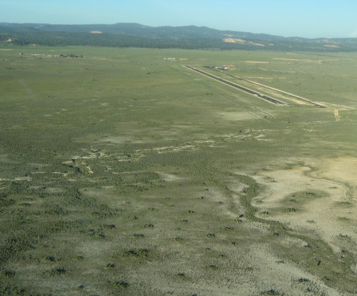 Aerial view of a vast, open landscape with sparse vegetation and a distant runway in the background.