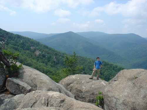A person stands on large rocks overlooking a lush green mountain landscape under a cloudy sky.