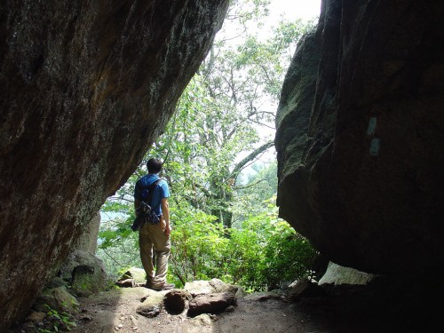A hiker stands between two large rocks, looking out at a lush, green landscape filled with trees.