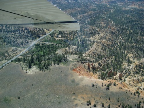 Aerial view of rugged terrain with trees, rocky formations, and a winding road beneath a plane's wing.