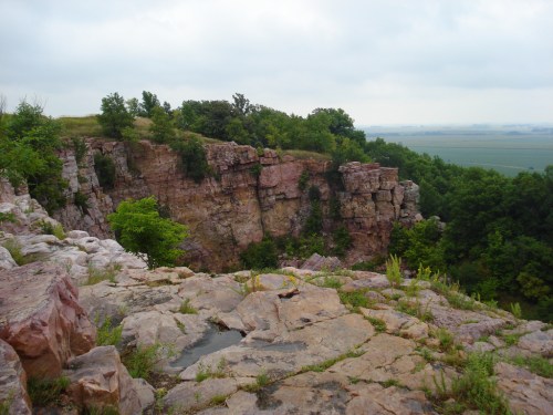 A rocky cliff with greenery on top, overlooking a vast landscape under a cloudy sky.