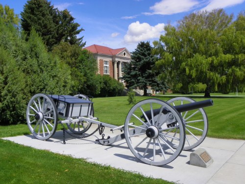 A historic cannon displayed on a grassy area, with trees and a building in the background under a blue sky.
