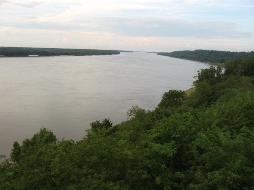 A wide river flows through lush green banks under a cloudy sky, surrounded by trees and distant hills.