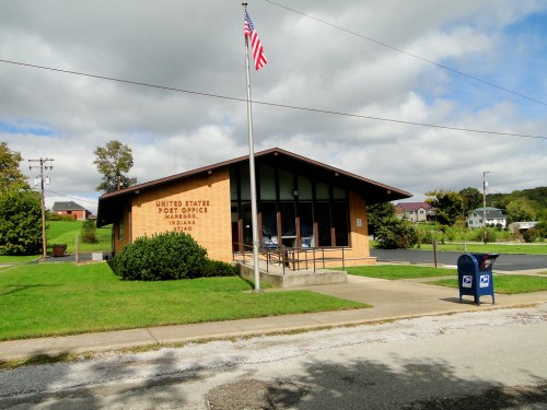 A small U.S. post office building with a flag, surrounded by green grass and a mailbox in front.
