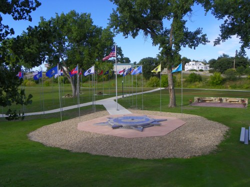 A park with a circular stone monument surrounded by flags and trees, featuring a pathway and green grass.
