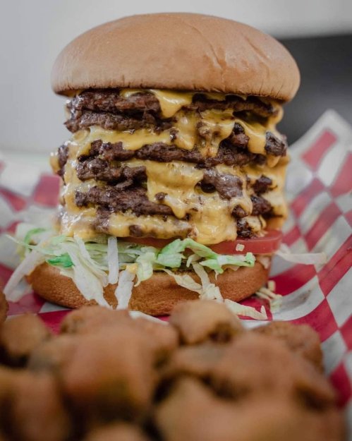 A towering burger with multiple beef patties and melted cheese, served on a bun with lettuce and tomato, alongside fried snacks.