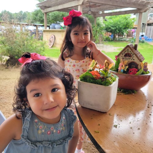 Two young girls with bows in their hair smile near a colorful garden decoration on a table outdoors.