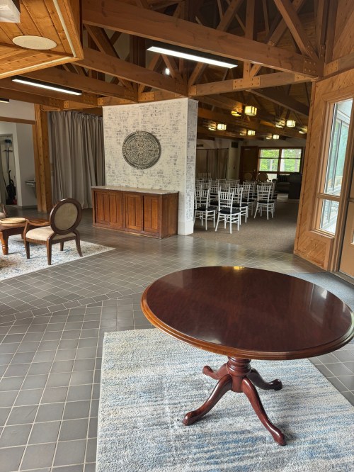 A spacious interior with wooden beams, a round table, and a seating area, leading to a room with white chairs.
