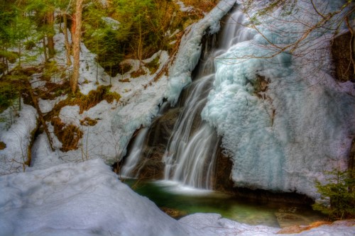A serene waterfall cascading over icy rocks, surrounded by snow and trees in a tranquil winter landscape.