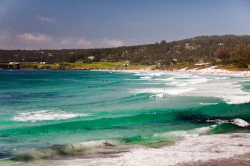 A scenic beach view with turquoise waves, surfers in the water, and a lush green hillside in the background.