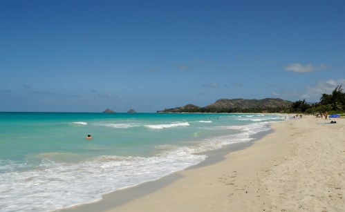 A serene beach scene with turquoise waters, gentle waves, and distant islands under a clear blue sky.