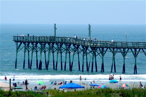 A wooden pier extends over the ocean, with beachgoers and colorful umbrellas on the sandy shore below.