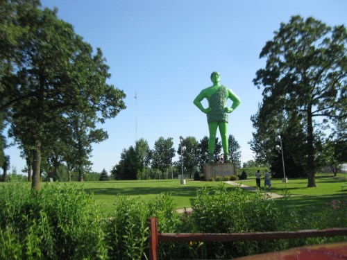 A large green statue of a man stands in a park, surrounded by trees and grass under a clear blue sky.