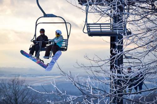 Two skiers on a chairlift, surrounded by frosty trees and a scenic winter landscape.