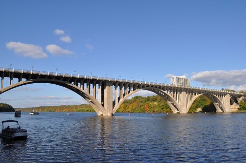 A concrete bridge spans over a river, with boats below and trees in the background under a clear blue sky.