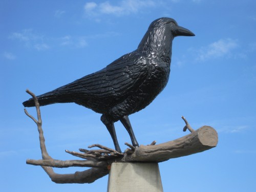 A large black raven sculpture perched on a branch against a blue sky.