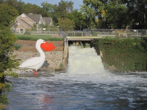 A large white duck statue stands near a waterfall, with greenery and houses in the background.