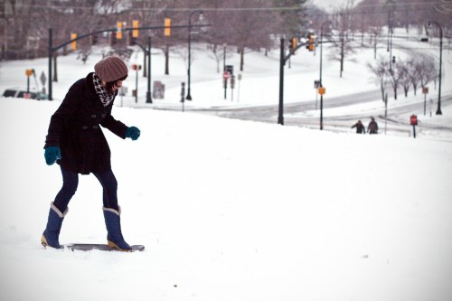 A person in winter attire walks through a snowy landscape, with a road and traffic signals in the background.