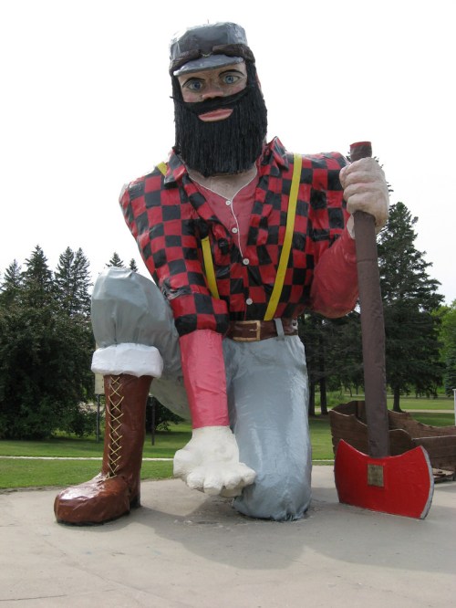 A large statue of a lumberjack in a plaid shirt, kneeling with an axe, surrounded by trees in a park setting.