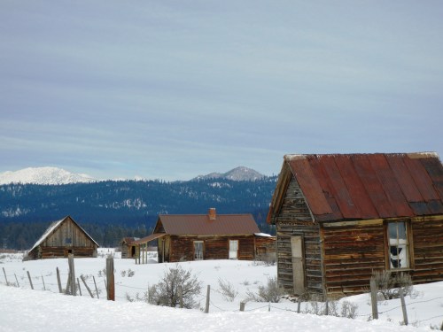 Abandoned wooden cabins with rusty roofs in a snowy landscape, surrounded by mountains under a cloudy sky.