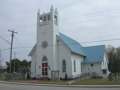 A white church with a blue roof and a tall bell tower, surrounded by a cemetery and power lines.
