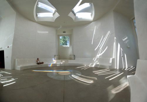 A spacious, minimalist room with white walls, skylights, and sunlight creating patterns on the floor. A person sits quietly.