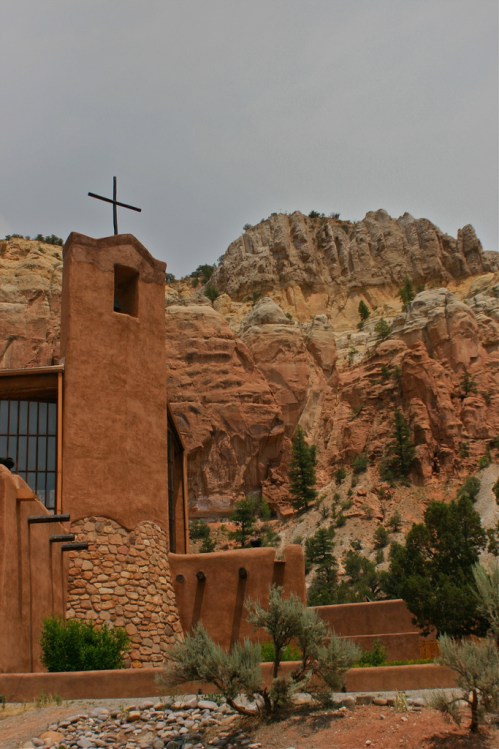 A rustic adobe church with a cross, set against a backdrop of colorful rocky cliffs and greenery.