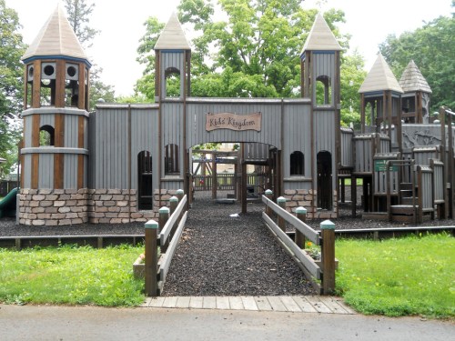 A large wooden playground with castle-like towers, surrounded by green grass and a wooden walkway.