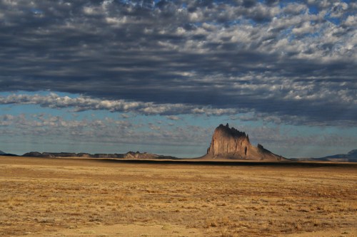 A dramatic rock formation rises from a vast, arid landscape under a cloudy sky.
