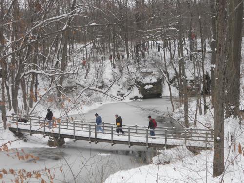 A snowy landscape with a wooden bridge over a frozen stream, where several people are walking through the winter scenery.