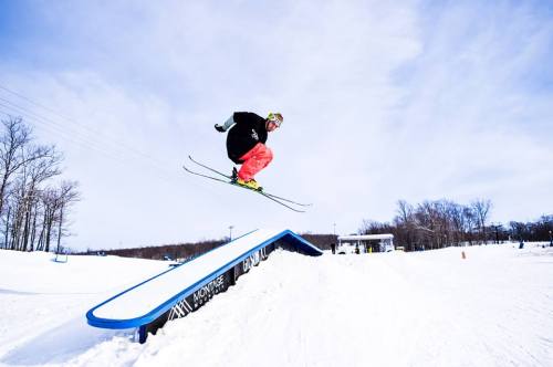 A skier performs a jump over a rail in a snowy landscape, showcasing athleticism against a clear sky.