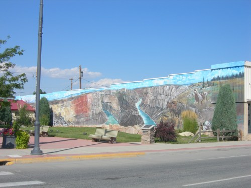 A colorful mural depicting a river and wildlife on a building, with trees and benches in a sunny outdoor setting.