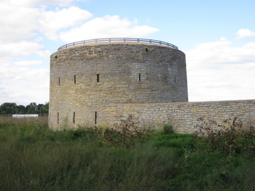 A round stone tower with a flat roof, surrounded by tall grass and a clear blue sky with scattered clouds.