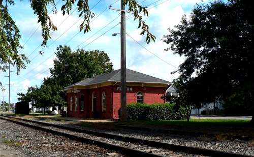 A red brick train station building beside railway tracks, surrounded by trees and a clear blue sky.