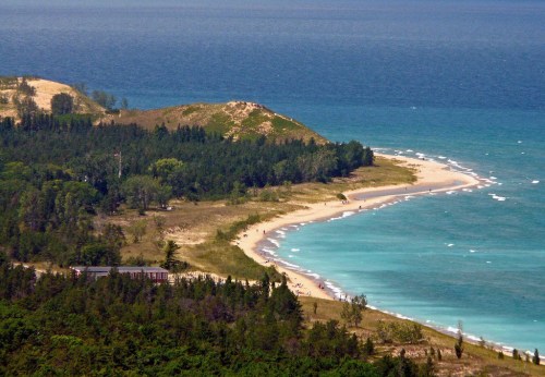 A scenic view of a sandy beach curving along a turquoise lake, surrounded by lush greenery and distant dunes.