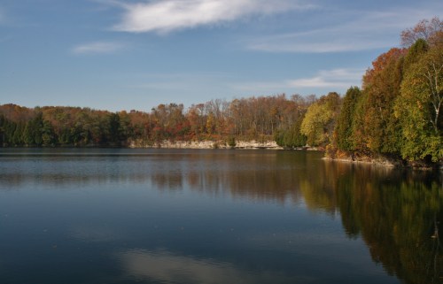 A serene lake surrounded by trees with autumn foliage, reflecting the clear blue sky and gentle clouds.