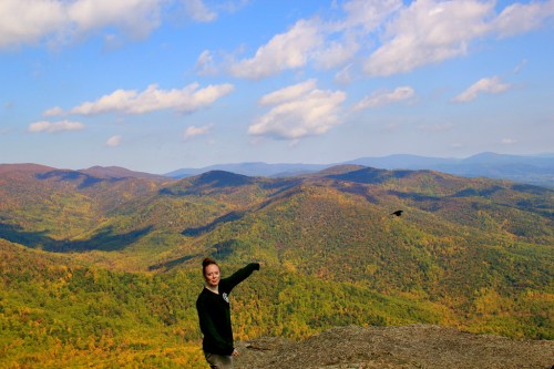 A person stands on a rocky outcrop, pointing towards a scenic view of colorful autumn mountains under a blue sky.