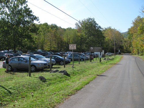 A parking lot filled with cars beside a road, surrounded by trees and a sign indicating a nearby park entrance.
