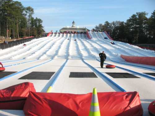 A snowy tubing hill with multiple lanes, red inflatable tubes, and a building in the background under a clear blue sky.