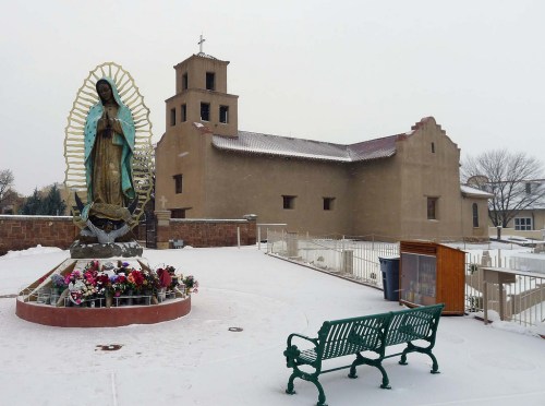 A snow-covered church with a statue of the Virgin Mary surrounded by flowers and a green bench in the foreground.