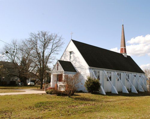 A white church with a tall steeple, surrounded by grass and trees, under a clear blue sky.
